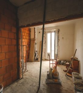 Gutted room mid-renovation with exposed brick walls, bare framing, and construction materials on the floor