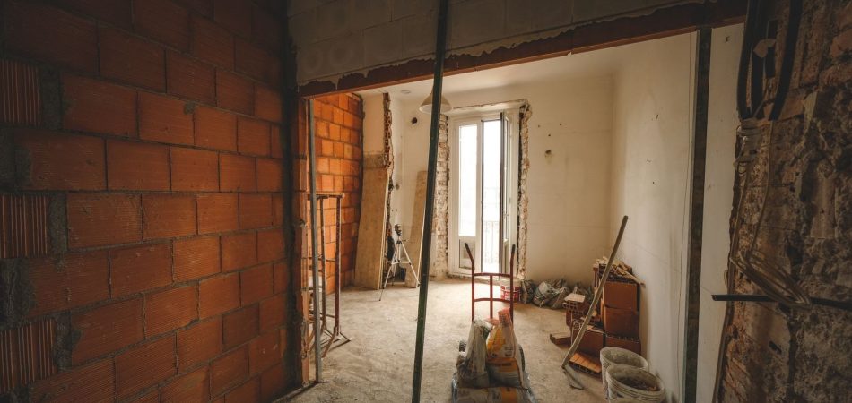 Gutted room mid-renovation with exposed brick walls, bare framing, and construction materials on the floor